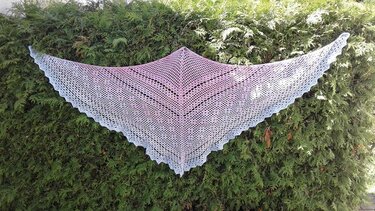 Gehäkeltes dreieckiges Spitzentuch in Pastellrosa bis Hellblau mit Muschelrand, gespannt vor einer Hecke