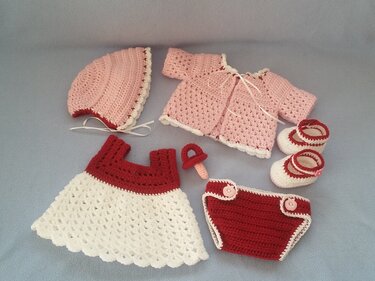 Handgehäkeltes Babykleidungsset in Rosa, Rot und Weiß mit Mütze, Jäckchen, Kleid, Windelhose, Schühchen und Schnuller.