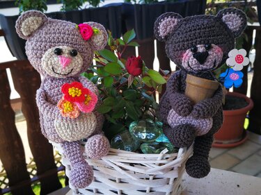 Zwei gehäkelte Amigurumi-Teddys (hellviolett und dunkelbraun) sitzen an einem weißen Korb neben einer roten Rose.