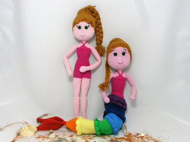 Zwei Amigurumi-Puppen auf einem Tisch mit Muscheln: eine im rosa Badeanzug, die andere mit bunter Meerjungfrauenflosse.