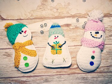 Drei einfache gehäkelte Schneemänner mit Mützen, Schals und Knöpfen auf hellem Holzbrett