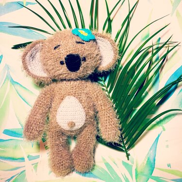 Gehäkelter brauner Amigurumi-Koala mit weißem Bauchteil und blauer Blume vor Palmenblatt