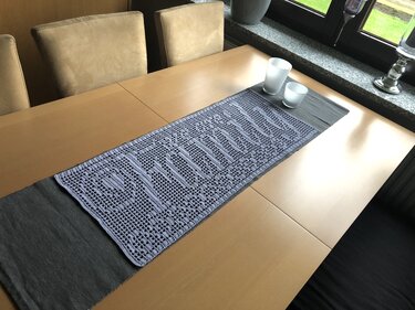 Gehäkelter lavendelgrauer Tischläufer mit geometrischem Gittermuster auf dunklem Unterläufer auf hellem Holztisch