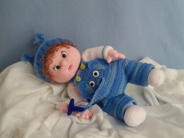 Gehäkelte Amigurumi-Babypuppe liegend in blauem Strampler und Mütze mit Schnuller