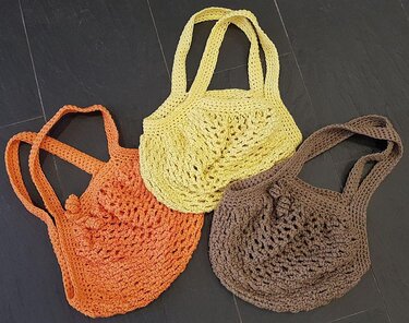 Drei gehäkelte Taschen in Gelb, Orange und Braun mit Netzmuster auf dunklem Untergrund.