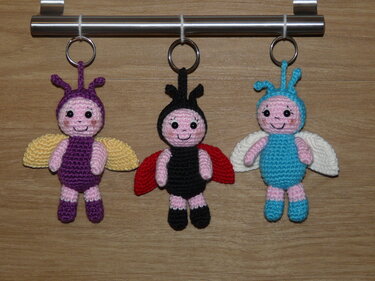 Drei gehäkelte Schlüsselanhänger in Käfer-/Insektenform (lila‑gelb, schwarz‑rot, blau‑weiß) an einer Metallstange.