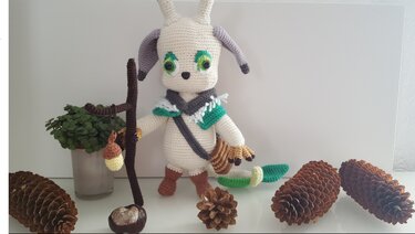 Weiß-beige gehäkelte Amigurumi-Figur mit Stab, Eichelanhänger und grünen Augen, umgeben von Tannenzapfen
