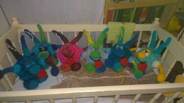 Fünf bunte, gehäkelte Amigurumi-Monster mit unterschiedlichen Gesichtern und Gliedmaßen liegen auf einer hellen Decke in einem weißen Kinderbett.