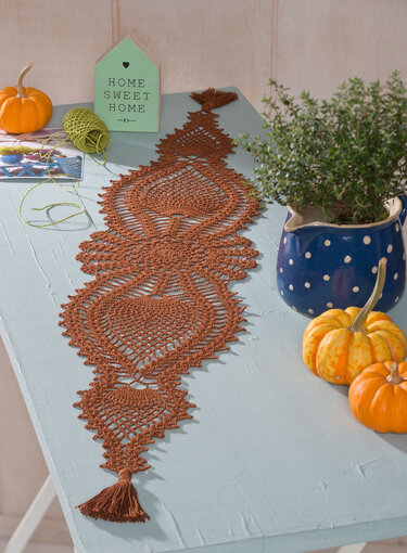 Rostbrauner gehäkelter Tischläufer mit Spitzenmuster und Quasten auf hellblauem Tisch, dekoriert mit Kürbissen und blauer Vase