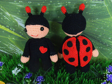 Zwei gehäkelte Marienkäfer-Amigurumi, eine von vorne mit Gesicht und Herz, die andere von hinten mit rotem, gepunktetem Panzer, auf grüner Wiese vor einem Blatt mit Tautropfen.