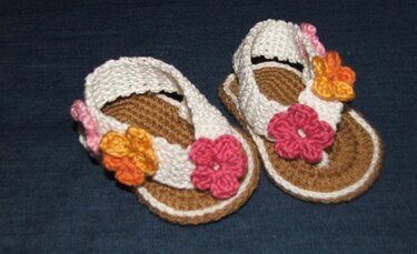 Gehäkelte Babysandalen mit brauner Sohle, weißen Riemen und rosa und orangefarbenen Blüten auf dunklem Untergrund.