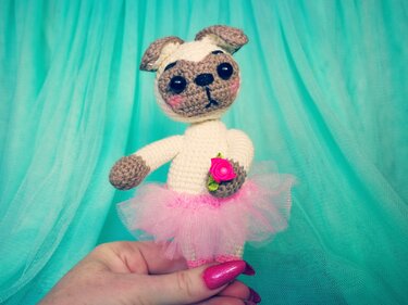 Gehäkelter Amigurumi-Mops mit beigem und braunem Gesicht, schwarzen Augen, pinkem Tüll-Tutu und kleiner pinker Rose, gehalten von einer Hand vor türkisfarbenem Hintergrund.