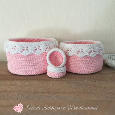 Drei gehäkelte Körbe in Rosa mit weißer Spitzenkante, zwei große und ein kleiner, nebeneinander auf einem Regal.