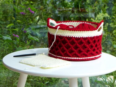 Red and white crocheted basket with textured stitch holding red yarn, with a crochet hook and white swatch on a white table and green foliage behind.