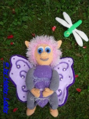 Amigurumi-Puppe mit pinkem Flauschhaar, lila Flügeln und blauen Augen neben einer kleinen grün-weißen gehäkelten Libelle auf einer grünen Wiese.