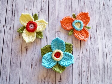 Drei gehäkelte Blumen-Applikationen in Gelb, Orange und Blau auf hellem Holzuntergrund