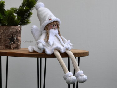 Weiße gehäkelte Amigurumi-Puppe mit Zipfelmütze, Schal, Flügeln und langen Beinen, sitzend auf einem Holztisch
