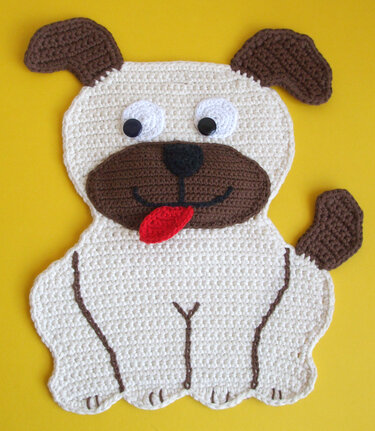 Gehäkelter Topflappen in Form eines sitzenden Hundes, cremefarben mit braunen Ohren, Schnauze und roter Zunge auf gelbem Hintergrund