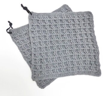 Zwei graue gehäkelte Topflappen mit Waffelmuster und schwarzen Aufhängeschlaufen auf hellem Hintergrund.