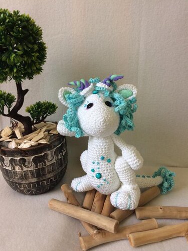 Weißer, handgehäkelter Amigurumi-Drache mit türkisfarbener Mähne und Perlen sitzt auf Holzstäbchen neben einer kleinen Pflanze.