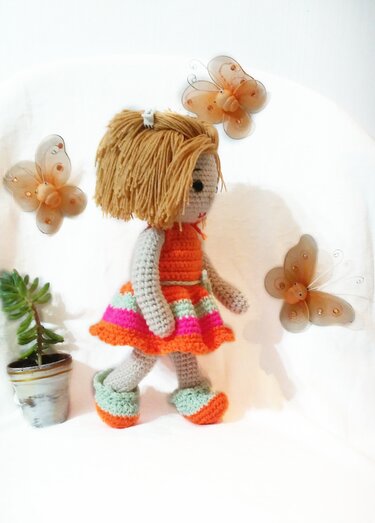 Crocheted amigurumi doll with light golden yarn hair wearing an orange top and a striped skirt in orange, pink and mint green, with matching booties — displayed with decorative butterflies and a small potted plant.