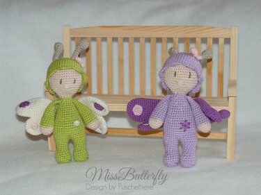 Zwei gehäkelte Amigurumi-Schmetterlingspuppen, eine grüne und eine lila, vor einer Holzbank.