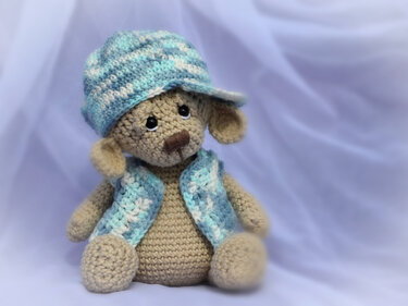 Beiger gehäkelter Teddybär mit blau-weiß gestreifter Mütze und Weste vor hellem Hintergrund