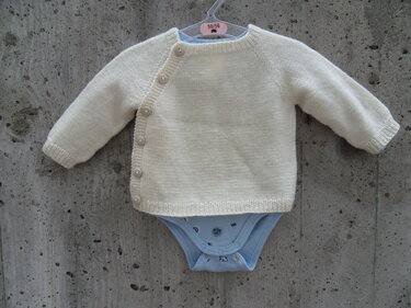 cremefarbene gestrickte Babyjacke mit seitlicher Knopfleiste und blauem Body, Größenlabel 50/56