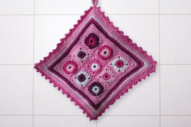 Gehäkelter quadratischer Topflappen mit neun Granny-Quadraten in Rosa- und Lilatönen, diagonal hängend an einer Fliesenwand