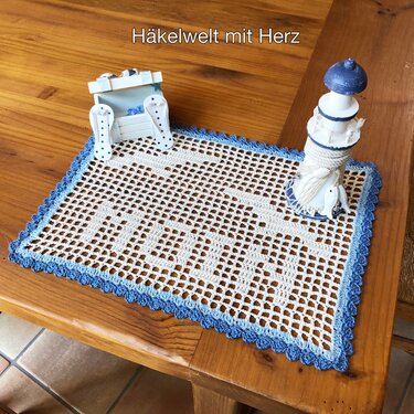 Cremeweißer gehäkelter Tischläufer mit blauem Wellenrand und Herzmuster auf einem Holztisch, dekoriert mit einem kleinen Leuchtturm und einer Schatztruhe.