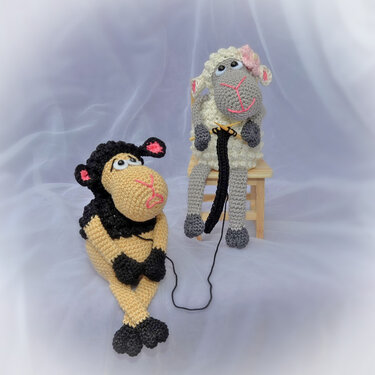 Zwei gehäkelte Amigurumi‑Schafe, liegend in Schwarz‑Beige und sitzend in Grau‑Weiß mit rosa Blüte