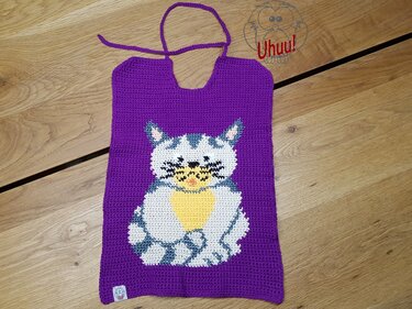 Lila gehäkeltes Babylätzchen mit eingehäkeltem Katzenmotiv und Bindeband, liegend auf Holz