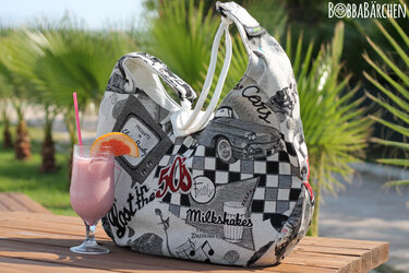 Große schwarz-weiße Stofftasche mit 50er-Jahre-Motiven und weißen Tragegriffen neben einem rosa Milkshake