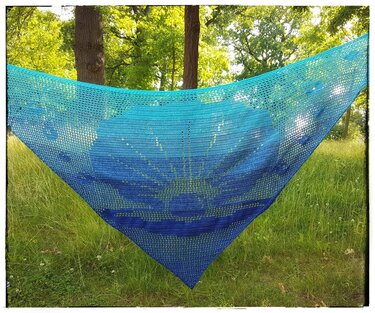 Türkis-blaues gehäkeltes Dreieckstuch mit Sonnenstrahlen-Motiv, im Grünen aufgehängt