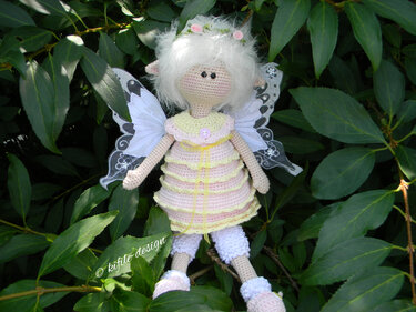 Gehäkelte Elfenpuppe mit weißem Haar, Flügeln und rosa-gelbem Kleid vor grünen Blättern