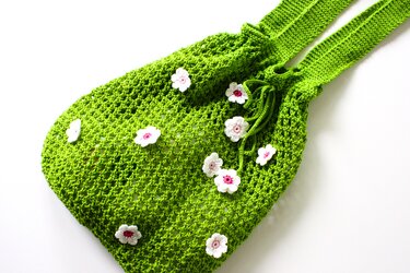 Gehäkelte grüne Beuteltasche mit Netzmuster, Kordelzug und weiß-rosa Blumen auf hellem Hintergrund
