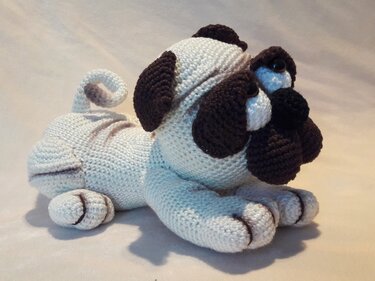 Cream and brown crocheted pug amigurumi lying down, showing curled tail and dark muzzle