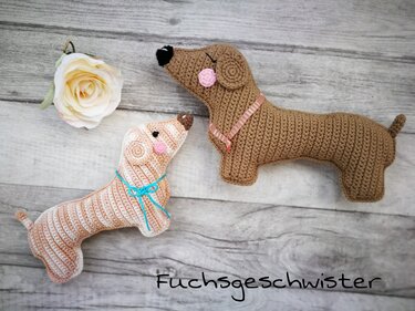 Zwei gehäkelte Dackel-Amigurumi auf hellem Holzuntergrund, einer gestreift mit blauem Band, der andere braun mit rosa Halsband