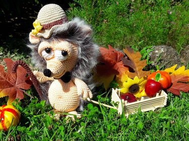 Amigurumi-Igel mit Hut und Häkelstock neben Mini-Wagen mit Kürbis und Herbstlaub.