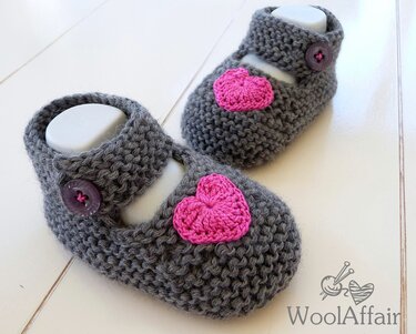 Graue gestrickte Babyschuhe mit rosa Häkelherz und Knopfriemchen auf hellem Untergrund