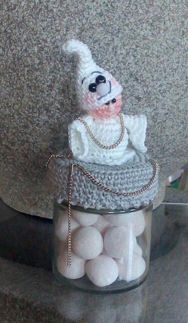 Gehäkelter Mini-Geist mit weißer Zipfelmütze und rosa Wangen auf grauem gehäkeltem Deckel über einem Glas mit weißen Kugeln, verziert mit einer dünnen Metallkette.