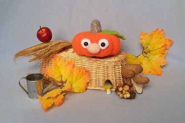 Gehäkelter Kürbis mit großen Augen auf einem geflochtenen Korb, umgeben von Herbstblättern, Pilzen und kleinem Apfel