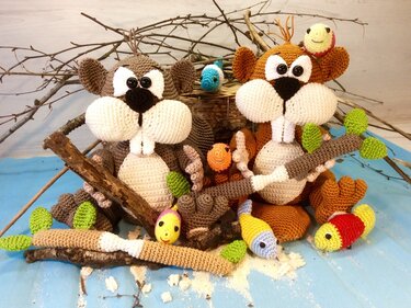 Zwei gehäkelte Amigurumi‑Nager in Braun und Beige mit großen Backen, sichtbaren Zähnen und kleinen gehäkelten Fischen vor Astdekor