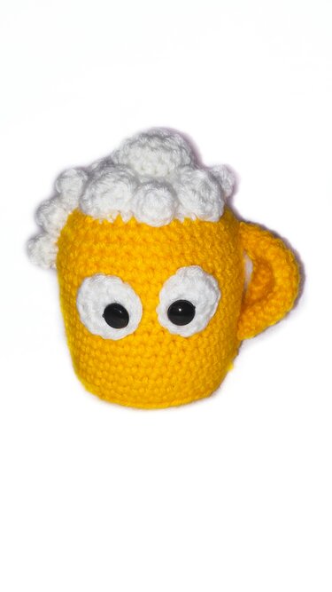Gehäkelter gelber Bierkrug-Amigurumi mit weißer Schaumkrone und großen schwarzen Augen