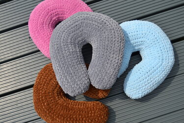 Vier U‑förmige gehäkelte Nackenkissen in Rosa, Grau, Hellblau und Braun auf grauen Terrassendielen.