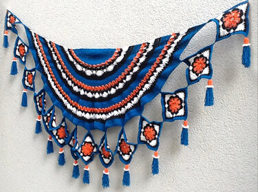 Blaues halbrundes gehäkeltes Tuch mit schwarzen, weißen und orangen Streifen; Rand aus Granny‑Squares mit blauen Quasten.