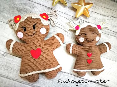 Nahaufnahme von zwei gehäkelten Lebkuchenfiguren mit weißen Zierstreifen, roten Herzen und Blumen auf hellem Holz