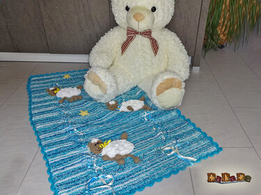 Gestrickte blaue Babydecke mit drei Schäfchen-Applikationen, Zierbändern und gezacktem Rand, darauf ein Teddybär