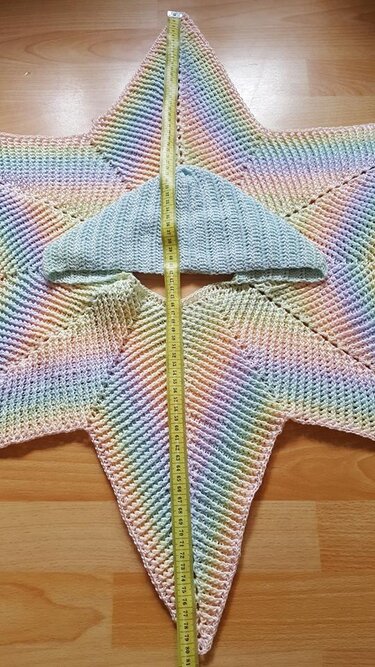 Gehäkelte sternförmige Baby-Wickeljacke in Pastellregenbogen mit hellblauer Mütze und Maßband auf Holzfußboden
