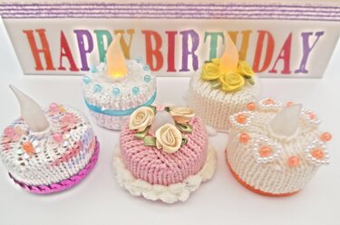 Nahaufnahme von fünf gestrickten Mini-Törtchen mit Rosen, Perlen und LED-Flämmchen vor buntem 'HAPPY BIRTHDAY'-Schriftzug.